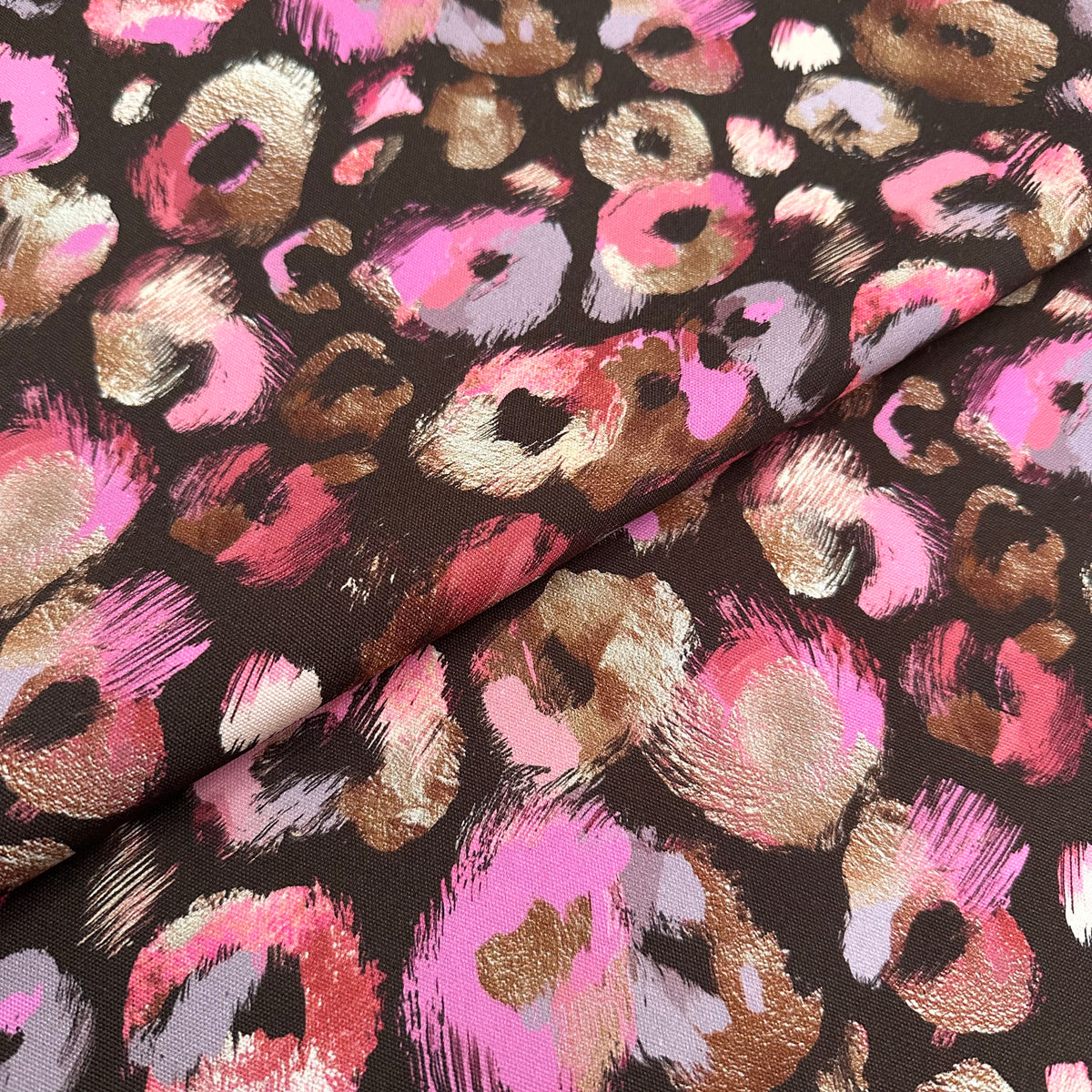 A close-up of a leopard print fabric featuring metallic pink, gold, and light pink spots on a dark brown background, with a soft fold in the material.